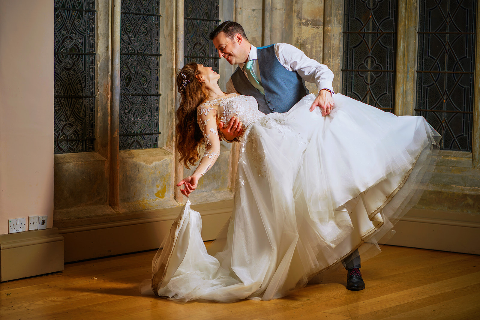 the groom leans his bride back after spinning her around at night with stained glass in the background as ware priory photographer, helen weir photography takes the shot.