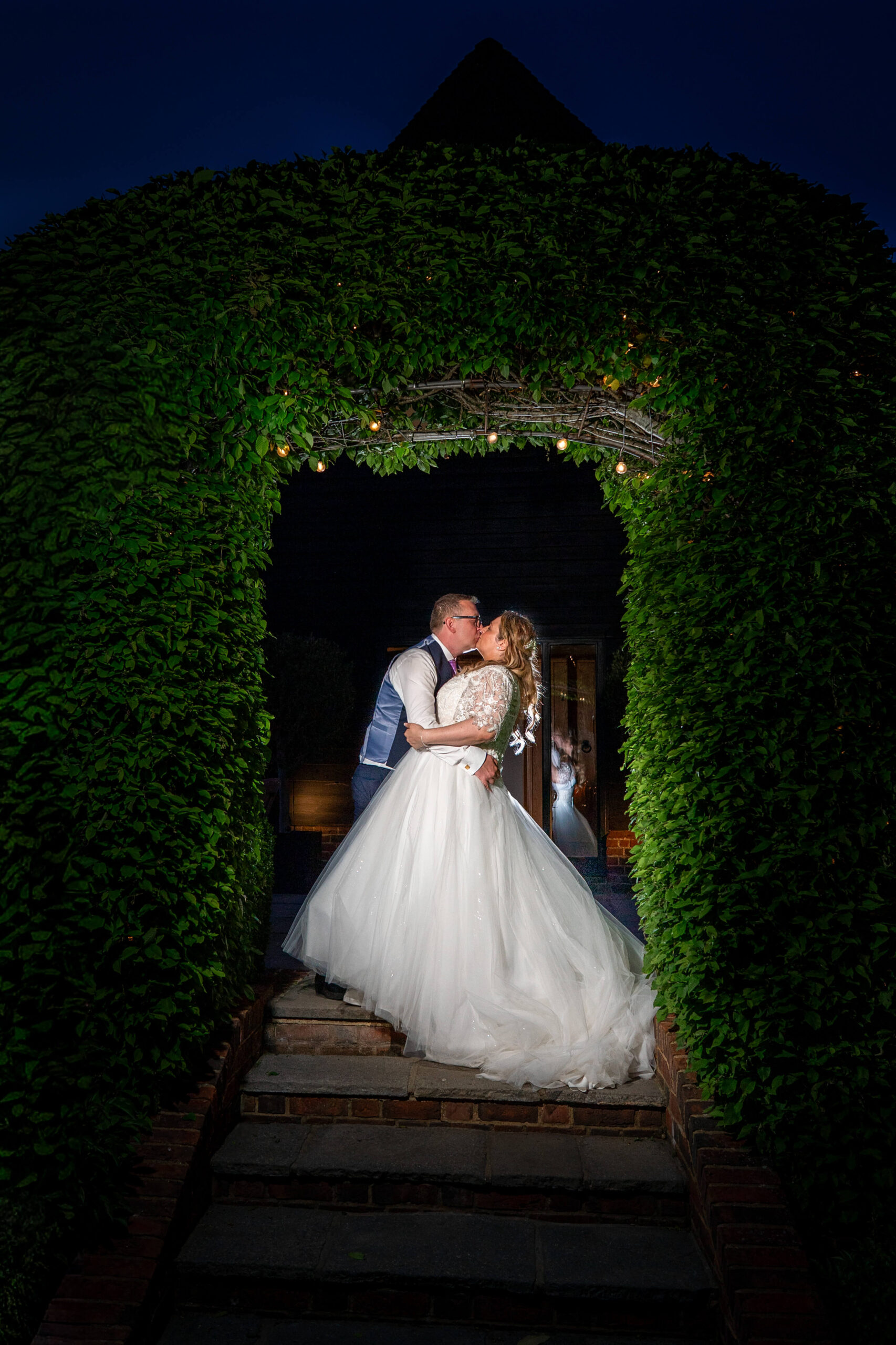 married later in life this bride and groom pose under a hedge archway at night