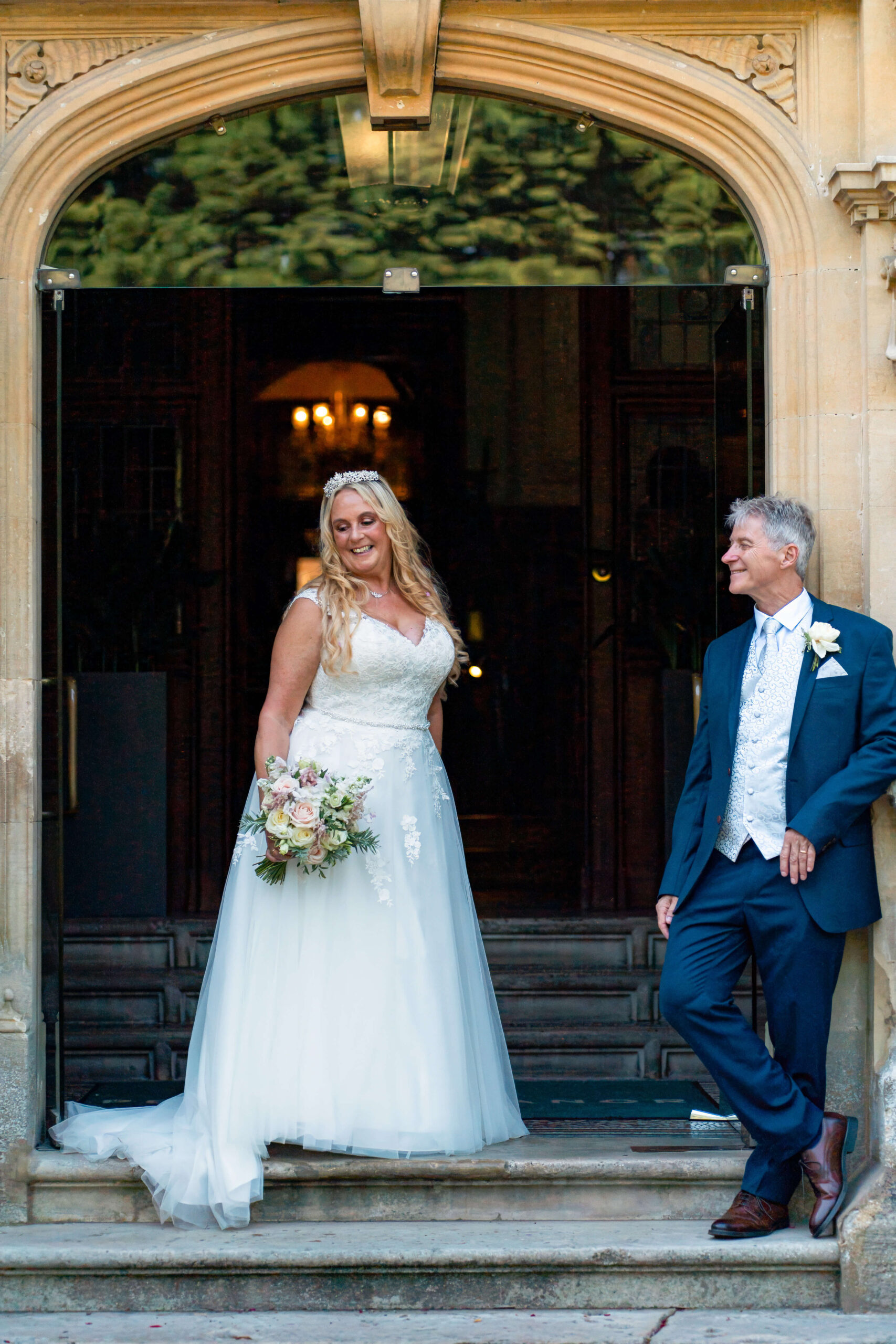 mature bride stands in door way whilst mature groom leans up against the wall looking at her